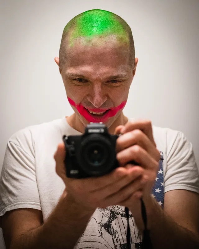 Self-portrait holding a camera, with green hair and red face paint.